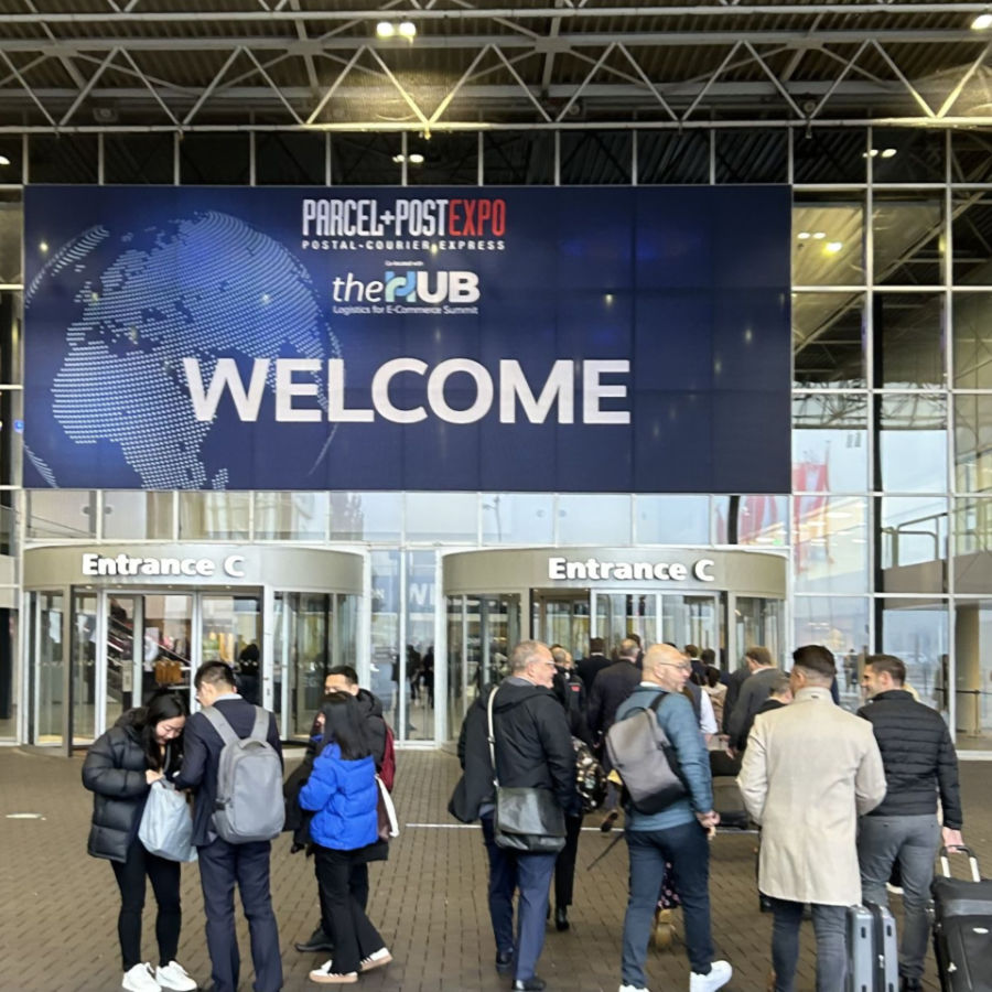 Entrance to the exhibition with the sign PARCEL+POST EXPO. Banner with a globe graphic. Entrance C doors. Group of people in front of the entrance.